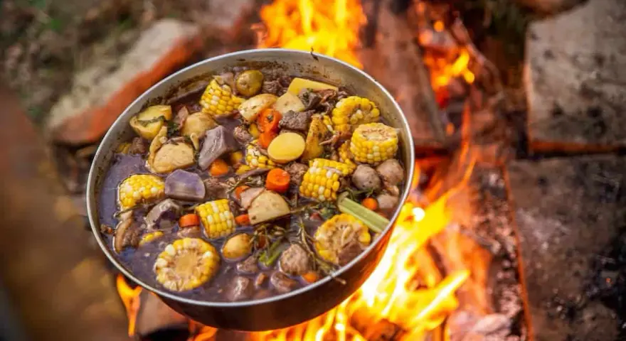 A pot of hearty camping meals, filled with corn, potatoes, carrots, and meat, simmers over a crackling campfire.