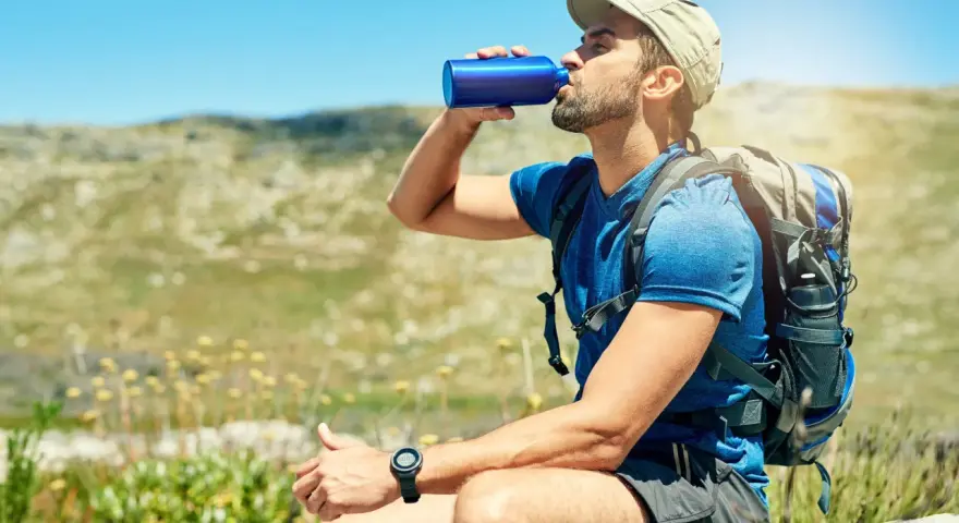A hiker takes a refreshing drink from a blue water bottle, demonstrating how to stay hydrated while hiking in a sunny, mountainous landscape.
