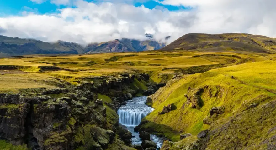 A winding path on the Laugavegur trail in Iceland leads past a mossy canyon with a cascading waterfall.