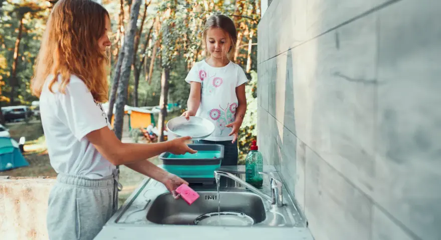 Two girls are washing dishes camping. One holds a pan over a sink while the other prepares to rinse it.