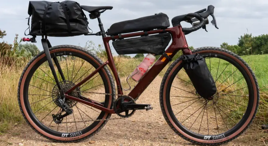 A maroon gravel bike with a full bikepacking setup, including saddle and frame bags, ready for an adventure on a dirt path.