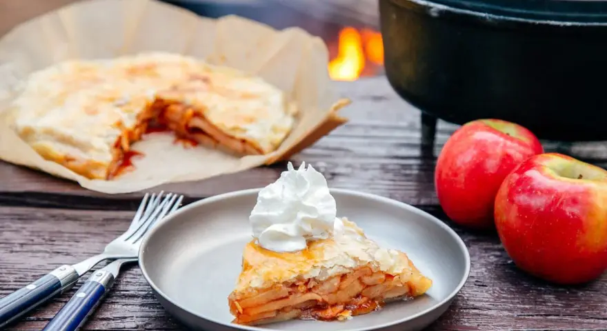 A slice of campfire apple pie, topped with whipped cream, sits on a plate next to fresh apples and a fork.