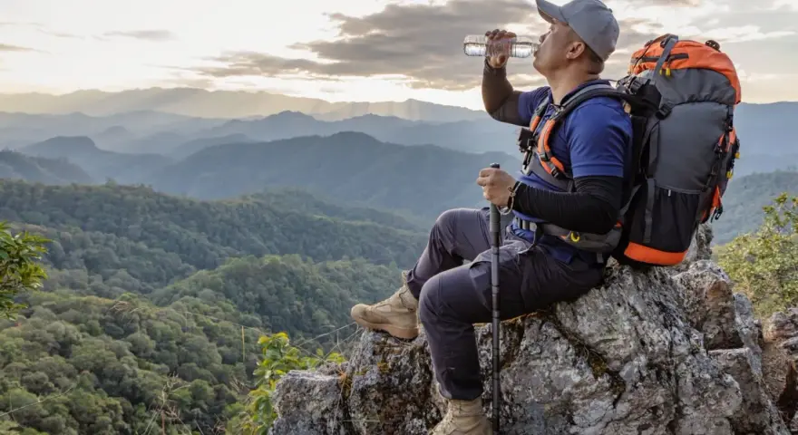 A hiker drinks water on a mountain peak, contemplating how much water to take backpacking.