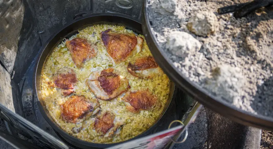 A delicious chicken and rice dish is cooking in a Dutch oven over a campfire, perfect for outdoor dutch oven campfire recipes.