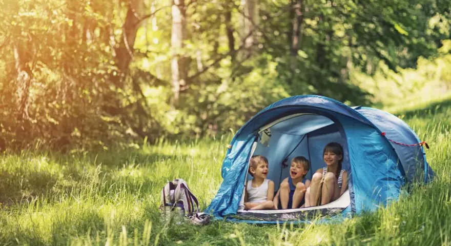 Three children laugh inside a blue tent, enjoying camping activities in a sun-dappled forest.