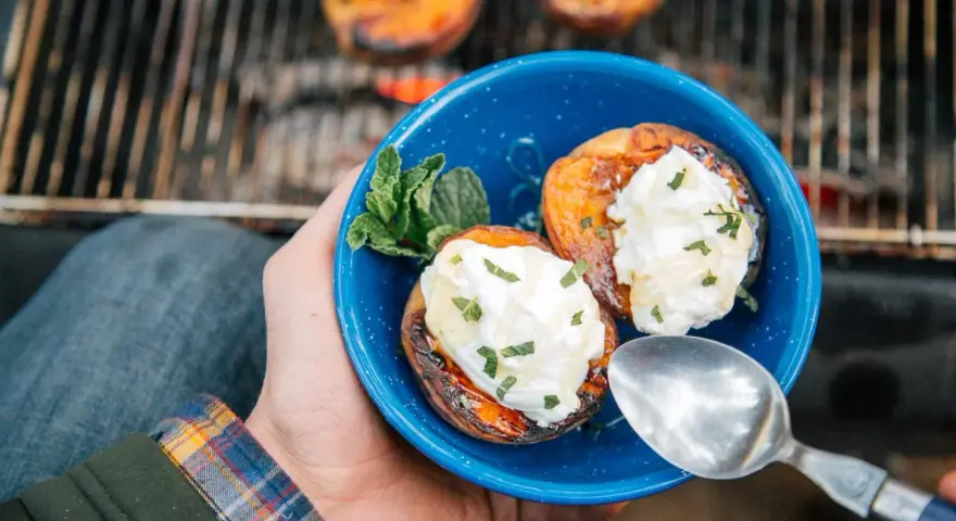 A hand holds a blue bowl with two grilled peaches topped with cream and mint. Campfire peaches are a perfect dessert.