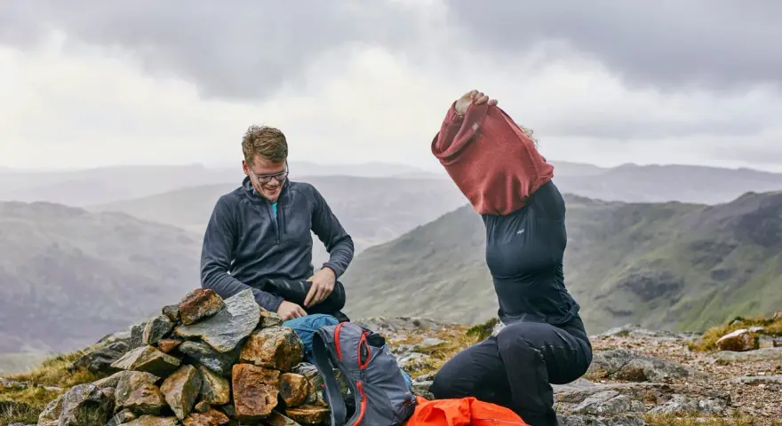 Hikers on a rocky summit adjust their backpacking clothes. One man smiles while sitting on a cairn, next to backpacks. Another person pulls on a red sweater.