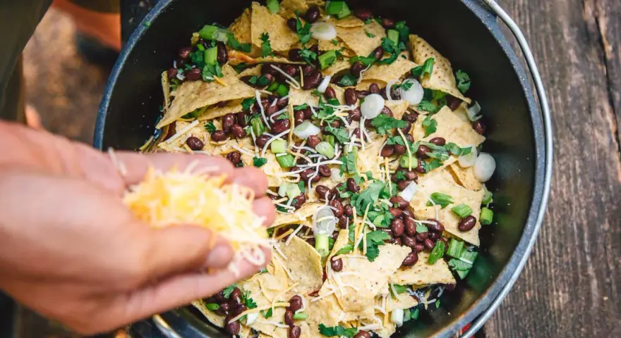 Making delicious camping nachos in a Dutch oven, adding shredded cheese to tortilla chips, black beans, and green onions.