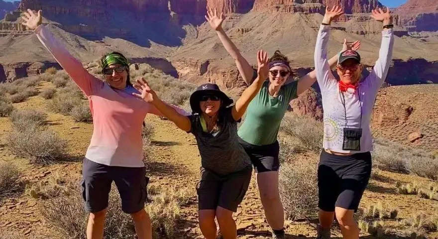 Four women pose with arms raised in a desert canyon, dressed for hiking in hot weather with shorts and light tops.