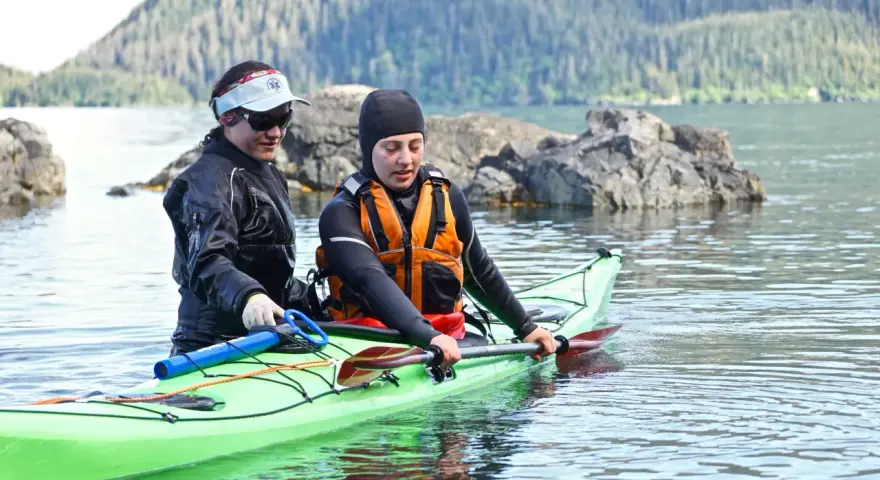 Two kayakers in a bright green kayak practice a paddle float re-entry in calm water, with a forested mountain in the background.