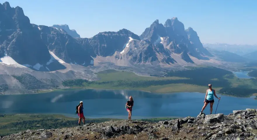 Three hikers enjoy a breathtaking view during their Tonquin Valley hike, with a stunning blue lake and jagged mountains behind them.