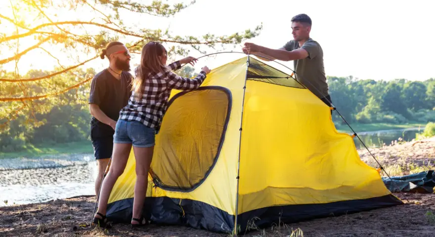 Friends setting up a bright yellow tent by a lake, learning how to choose a campsite with a beautiful sunset.