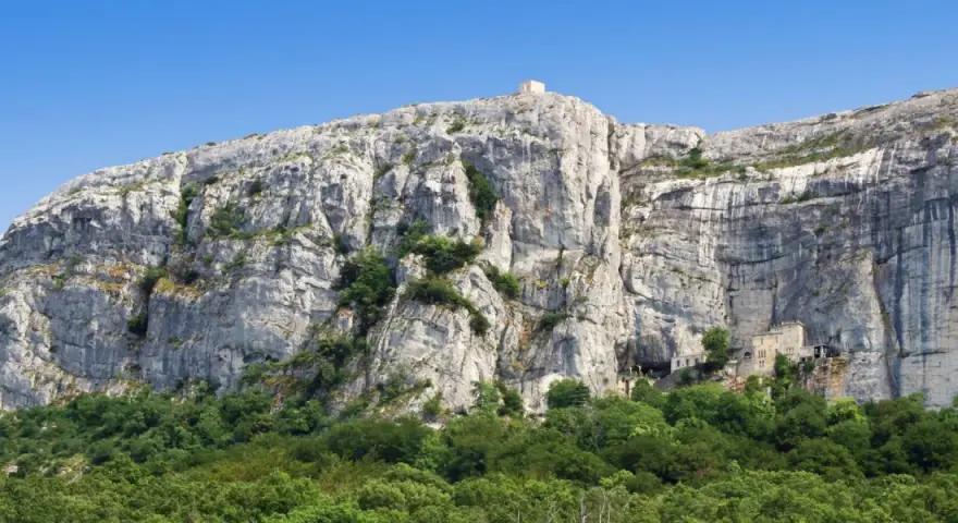 A majestic cliff face rises above a lush green forest, with a small building nestled into the rock at Sainte Baume.