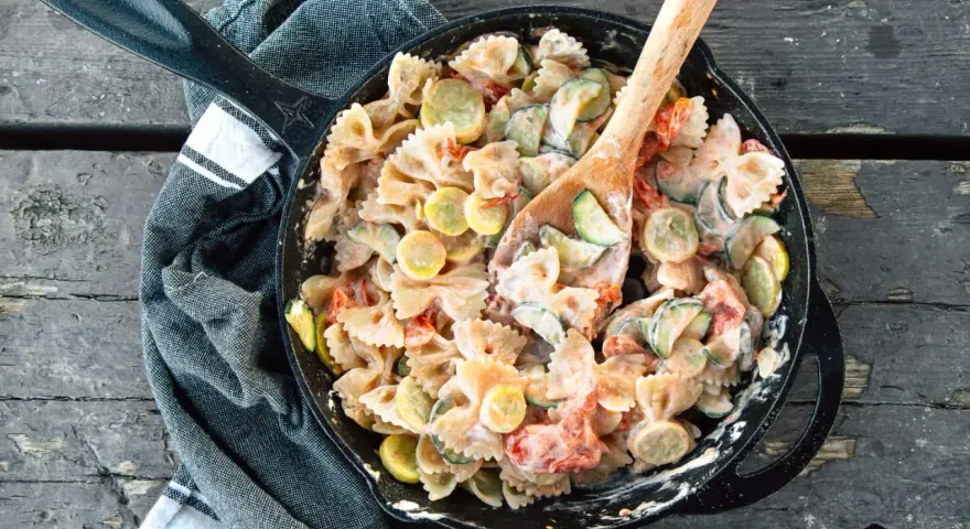 A hearty camping pasta dish with farfalle, zucchini, and tomatoes in a creamy sauce, served in a cast-iron skillet.