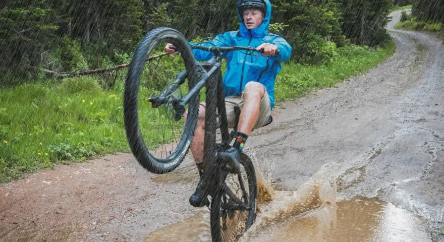 A mountain biker in a blue jacket performs a wheelie through a large puddle, splashing water as it rains heavily on a forest trail.