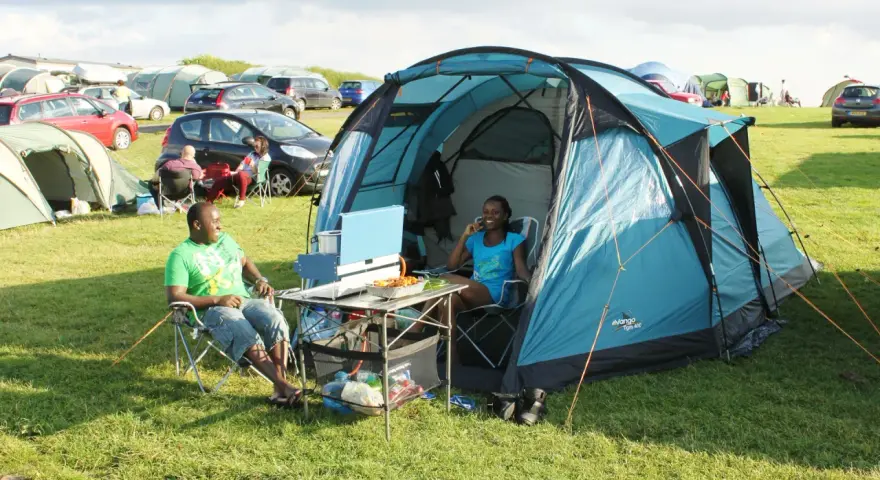 A couple enjoys a meal at their campsite, a perfect example of how to camp. They sit in chairs beside a blue tent, with food laid out on a portable table.