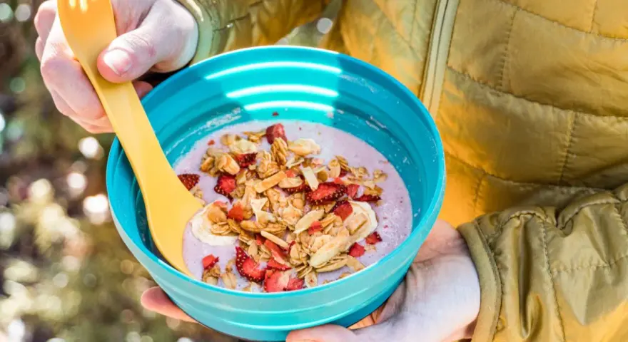 A person in a yellow jacket holds a blue bowl filled with a pink smoothie topped with granola, dried strawberries, and banana slices. Perfect for camping breakfast ideas.