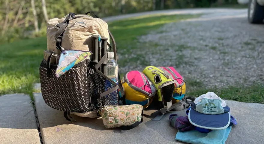 Ready for adventure! A large backpack, hiking poles, water bottle, and colorful pouches are laid out with camping gear, including a hat and gloves.