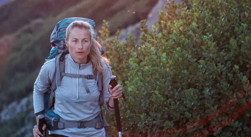 A woman with blonde hair, wearing a grey jacket and a large backpack, is backpacking hiking up a rocky, vegetated slope. She holds a trekking pole.