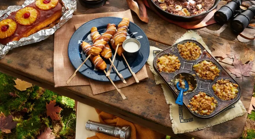 A spread of cozy camping food: pineapple upside-down cake, pigs in a blanket, apple crumble muffins, and a skillet brownie with ice cream.