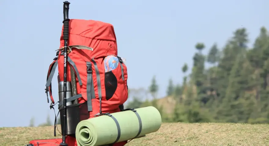 A red backpack with a rolled sleeping pad strapped to it, and trekking poles. This image shows how to pack a backpacking pack for an adventure.