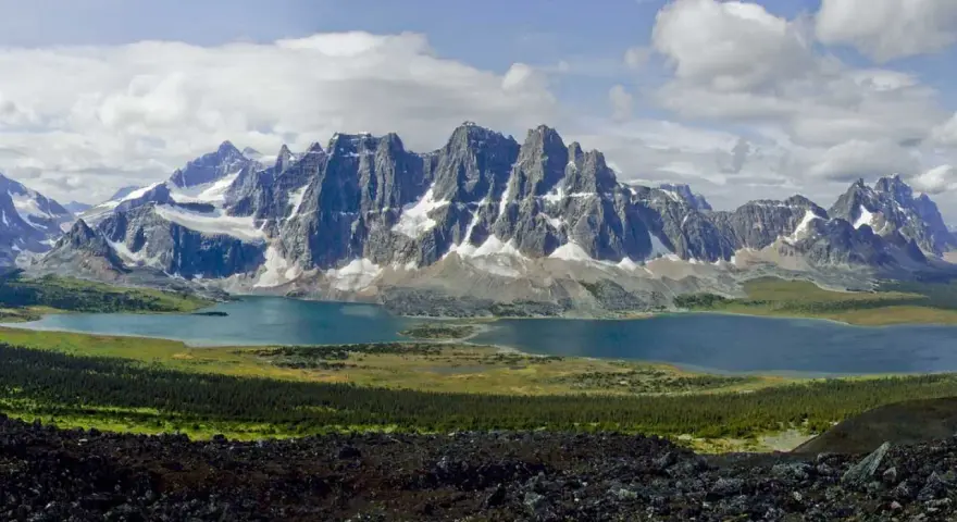A vast mountain range with snow-capped peaks overlooks a serene blue lake. The Tonquin Valley Trail offers this breathtaking view of rugged beauty.