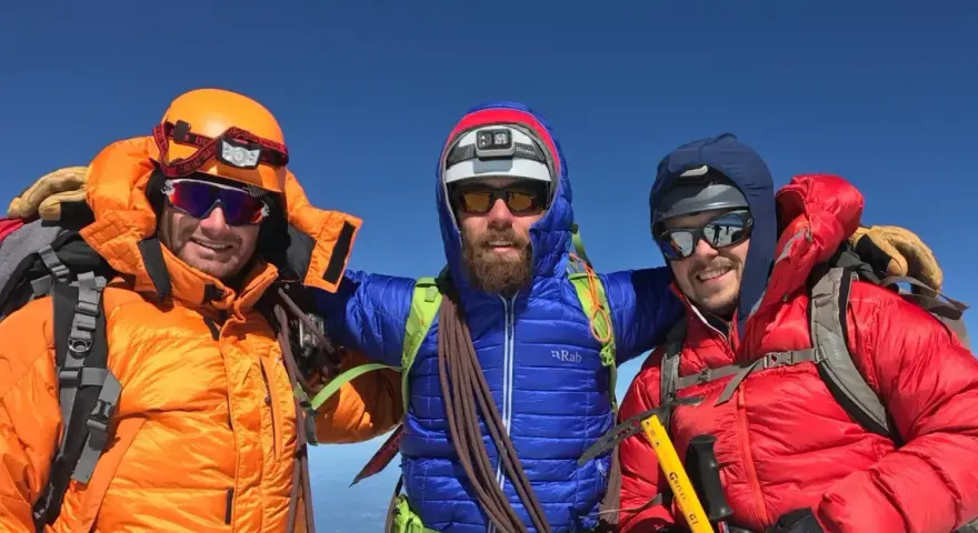 Three smiling mountaineers in bright down fill jackets and helmets pose on a sunny summit, ready for adventure.