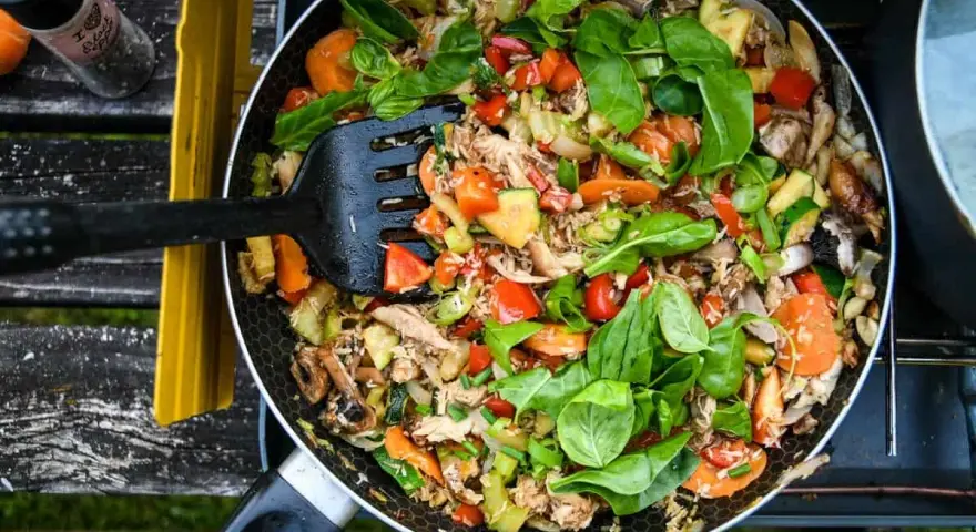 A colorful stir-fry with chicken, carrots, zucchini, and peppers, perfect for camp stove recipes. A black spatula is mixing the ingredients in a pan.