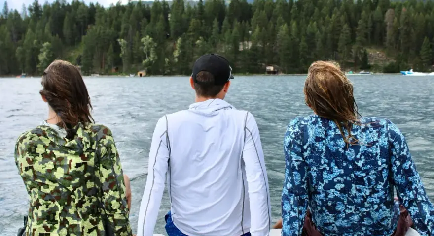 Three friends enjoy a lake view, wearing cooling clothing for extreme heat. The camouflage and blue patterned shirts offer sun protection.