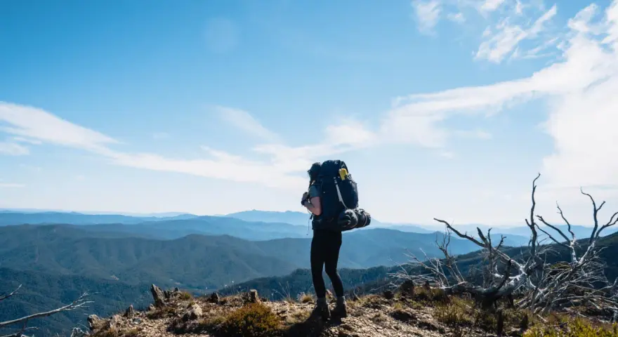 A lone hiker stands on a mountain ridge, ready for backpacking and hiking, with a vast expanse of blue mountains stretching to the horizon under a clear sky.
