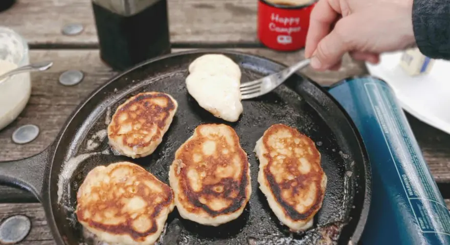 Frying pancakes in a cast iron skillet is a delicious answer to what to eat when camping. A fork lifts a fresh pancake, ready for syrup.