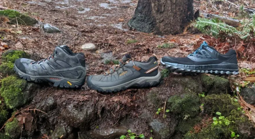 Three pairs of shoes on a wet forest floor: hiking boots, trail running shoes, and another pair of trail running shoes.