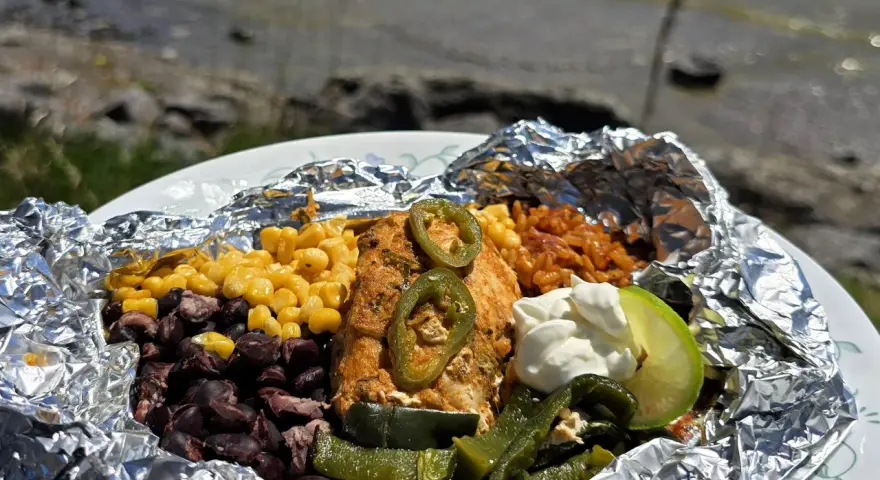 Delicious campfire chicken foil packets with corn, black beans, rice, jalapeños, and a lime wedge, ready to be enjoyed by the water.