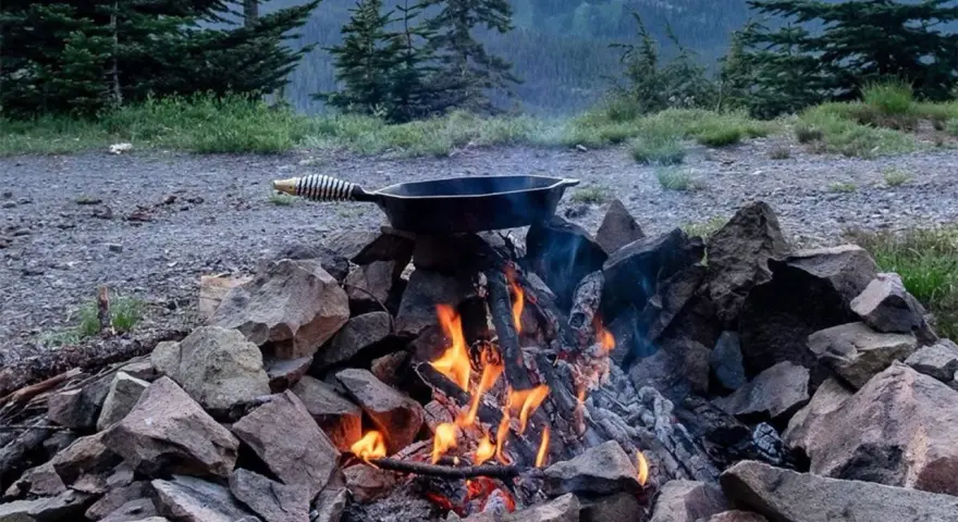 A cast iron skillet sits over a crackling campfire, ready for a delicious cast iron camping meal amidst a scenic mountain backdrop.