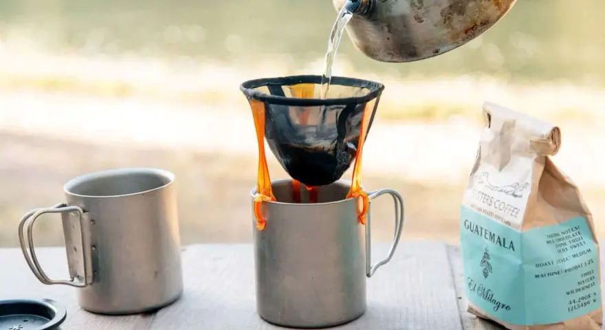 Making coffee while camping: hot water pours from a kettle into a pour-over filter, brewing coffee into a metal mug. A bag of Sisters Coffee Guatemala sits nearby.