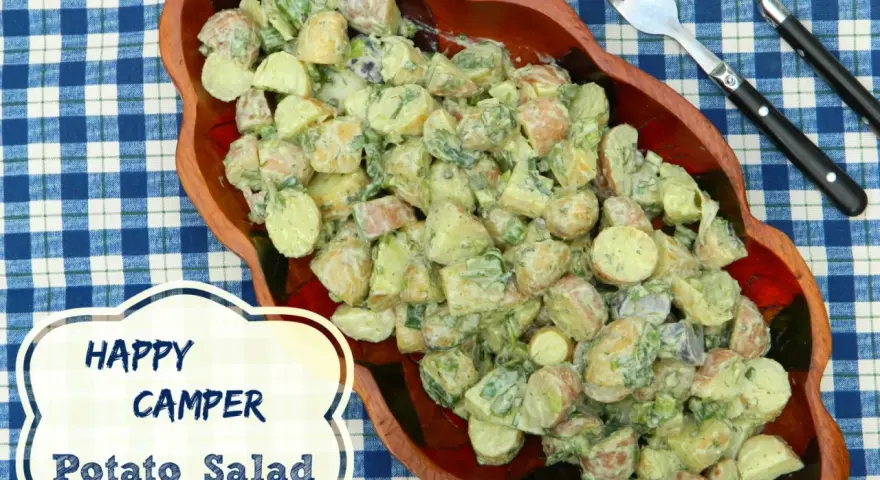 A hearty potato salad, perfect for happy campers, is served in a wooden bowl on a blue checkered tablecloth.