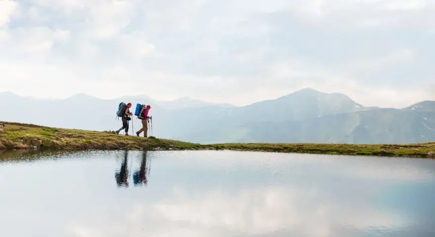 Two hikers with backpacks walk along a grassy ridge beside a calm lake, with misty mountains in the background. This image inspires thoughts on how to go backpacking.