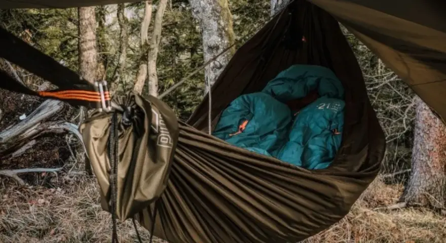 Cozy hammock camping setup in a forest. Two teal sleeping bags are nestled inside a brown hammock, ready for a night under the stars.