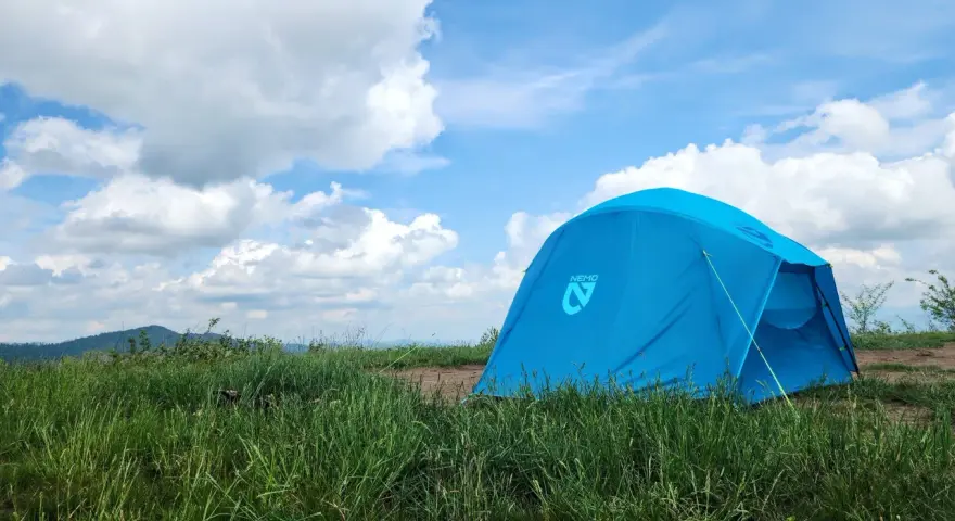 A bright blue Nemo tent stands in a grassy field under a cloudy sky. This Nemo Aurora tent is ready for adventure.