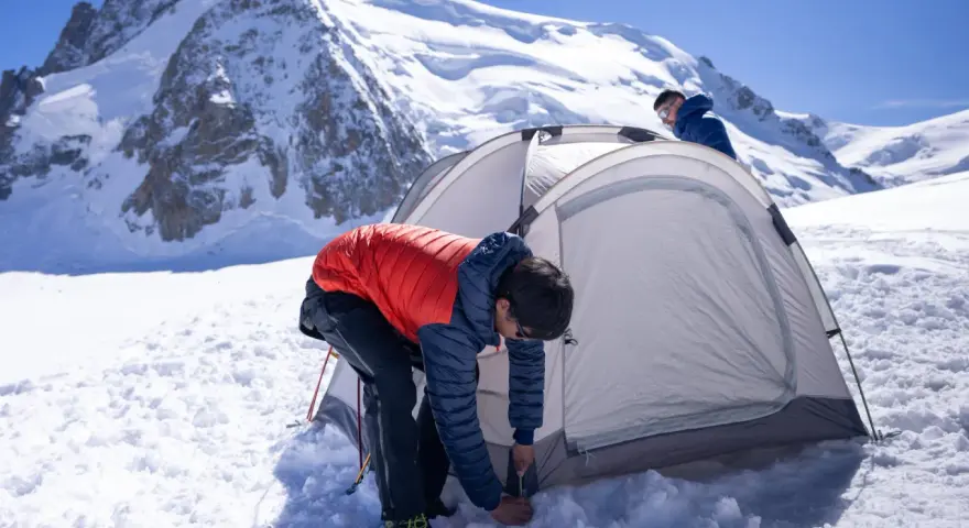 Two people set up a tent for winter camping amidst a snowy mountain landscape under a clear blue sky.