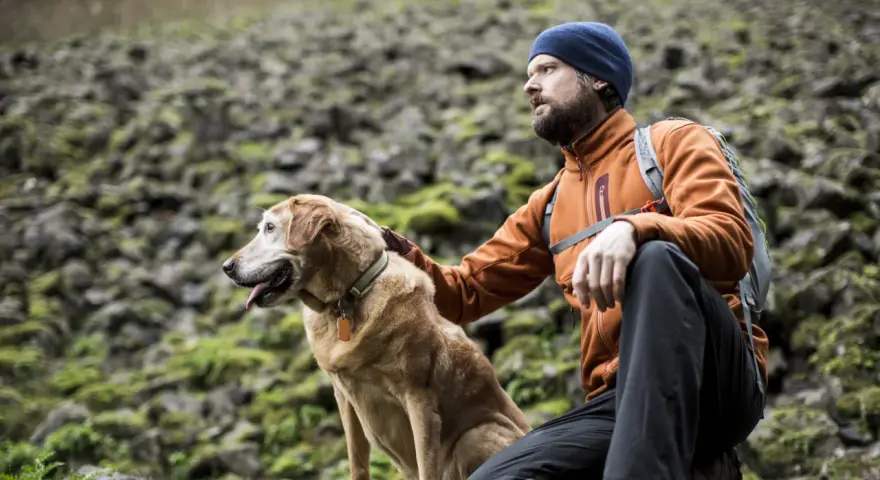 A man in an orange jacket and blue beanie rests during a hiking trip with his golden dog.