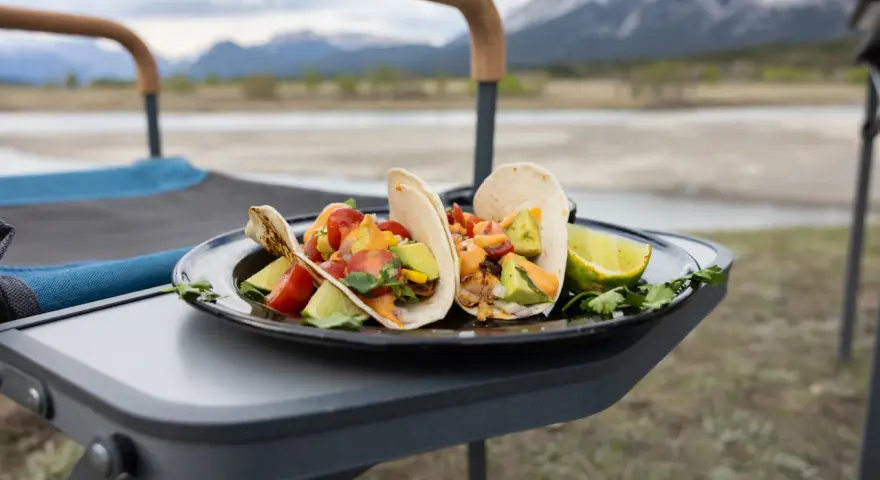 Delicious camping tacos with avocado, tomatoes, and lime, served on a plate on a portable table with mountains in the background.