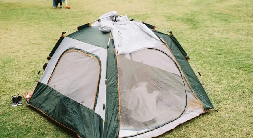 A green and gray tent is partially set up on a grassy field. A child plays in the background, learning how to set up a tent.