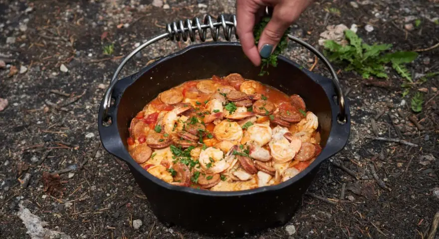 A hand garnishes a hearty dutch oven paella with fresh parsley. This campfire meal features shrimp, sausage, and rice in a rich tomato sauce.