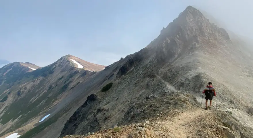 A hiker with a backpack and trekking poles walks along a narrow mountain ridge, a scenic path to learn how to get better at hiking.