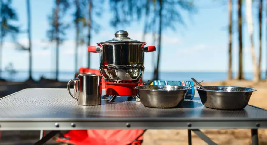A complete camping kitchen setup with a stove, pots, bowls, and mugs on a table, ready for outdoor cooking by a lake.