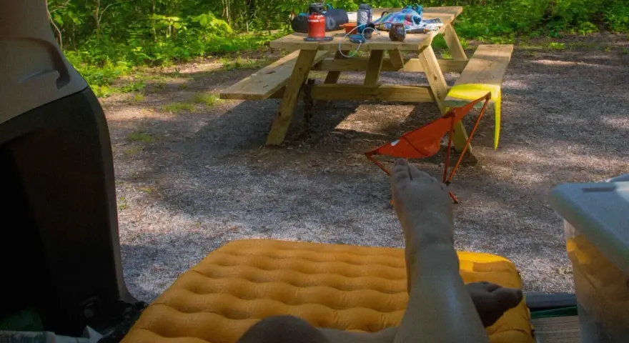 A person's legs rest on an orange sleeping pad inside a car. A picnic table with camping gear sits in a wooded area, ready for a road trip checklist.
