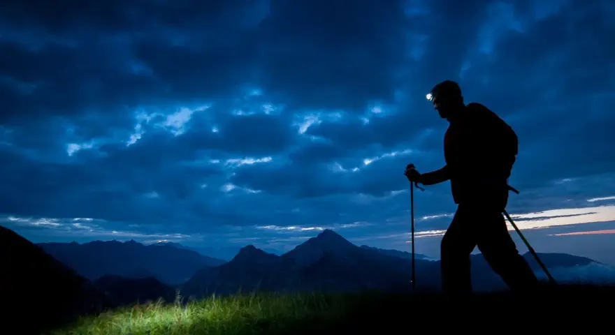 Silhouette of a hiker with a headlamp on, trekking poles in hand, on a night hike through a dramatic, cloudy sky over mountains.