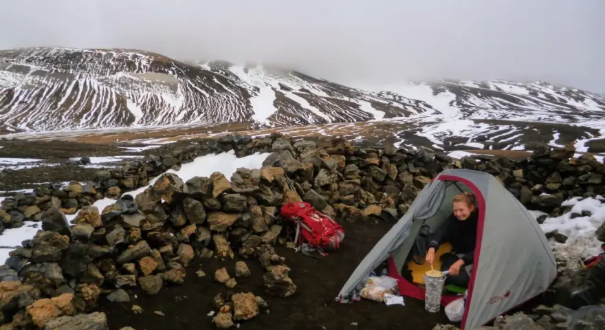 A hiker prepares a meal inside a tent at one of the Laugavegur trail huts, surrounded by snow-dusted mountains and a stone wall.