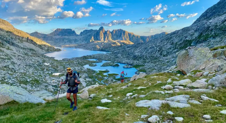 Hikers ascend a grassy slope on the HRP trail, with a stunning mountain lake and jagged peaks in the background under a cloudy sky.